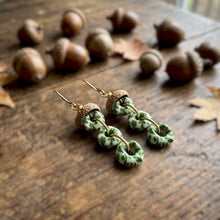Load image into Gallery viewer, Dangle earrings with green and white spotted bead-like drops and brown cork-look caps, photographed on a wooden surface with scattered acorns and leaves in the background.