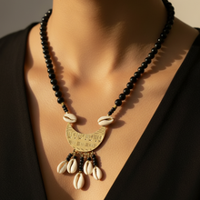 Load image into Gallery viewer, A close-up of a model wearing a black beaded necklace with a gold-toned crescent pendant engraved with script, accented by white cowrie shells.