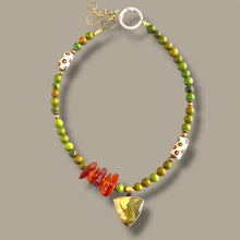 Load image into Gallery viewer, A beaded necklace with green and yellow beads, orange-red agate chips, white beads with gold dots, and a gold shield-shaped pendant, displayed on a neutral gray background.