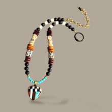 Load image into Gallery viewer, A beaded necklace with a geometric heart-shaped pendant, featuring black beads, turquoise, white, brown, and gold-toned elements, displayed on a solid taupe background.