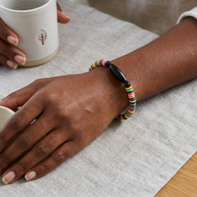 Load image into Gallery viewer, A multicolor beaded bracelet with a black central bead worn on a person's wrist, resting on a light gray linen cloth with hands holding a white ceramic mug in the background.