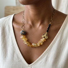 Load image into Gallery viewer, A close-up of a person wearing a bohemian-style statement necklace. The necklace features a strand of irregular yellow and amber beads, a black beaded section, a textured black and gold bead, and gold-toned metal links and charms, worn over a cream-colored linen top.