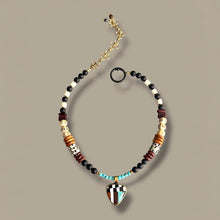 Load image into Gallery viewer, A beaded necklace with a guitar-shaped pendant, featuring black, white, brown, and turquoise beads, displayed on a neutral taupe background.