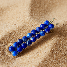 Load image into Gallery viewer, A brass hair barrettes embellished with faceted lapis sitting on sand. 