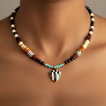 Load image into Gallery viewer, A beaded necklace with a geometric inlay pendant, shown on a model's neck. The necklace features black and white beads, turquoise blue beads, patterned wooden beads, and a central pendant with a black, white, orange, and turquoise geometric design in a gold-tone frame.