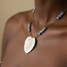 Load image into Gallery viewer, A close-up of a model wearing a necklace with a large white leaf-shaped pendant and blue-and-gold beaded accents on a gold-toned chain, against a warm beige background.