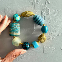 Load image into Gallery viewer, Teal, black, and brass beaded bracelet held by a hand on a textured light gray background