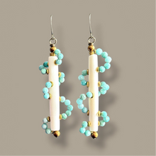 Load image into Gallery viewer, A pair of long dangle earrings featuring vertical white cylindrical beads wrapped with strands of small turquoise-blue beads and accented with gold-toned spacers and hooks, set against a solid taupe background.