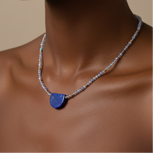 Load image into Gallery viewer, A close-up of a model wearing a necklace with a small, polished blue pendant and a delicate silver beaded chain against a warm beige background.