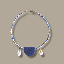 Load image into Gallery viewer, A sodalite beaded necklace featuring a large, polished blue semicircle pendant at the center, flanked by two small white cowrie shells and accented with blue, white, and brass beads, set against a solid taupe background.