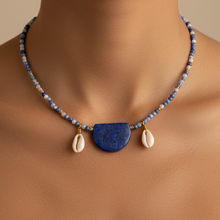 Load image into Gallery viewer, A close-up of a model wearing a beaded necklace with a blue semicircle pendant and two white cowrie shells, set against a warm beige background.