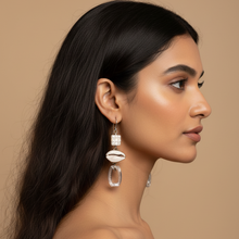 Load image into Gallery viewer, A woman in profile wearing a long silver-toned drop earring with a white shell-shaped charm and clear teardrop pendant, against a warm beige studio background.