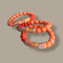 Load image into Gallery viewer, A set of three beaded bracelets with orange and red beads, featuring two large patterned orange and yellow geometric beads and one smaller orange and yellow geometric bead, displayed on a solid taupe background.