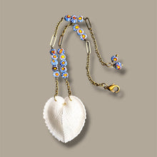 Load image into Gallery viewer, A heart-shaped white shell pendant necklace with a brass chain decorated with blue, yellow, and white daisy glass beads, shown on a solid taupe background.