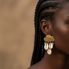 Load image into Gallery viewer, Close-up of a woman wearing a gold cloud-shaped earring with cowrie shells and turquoise beads against a blurred background.