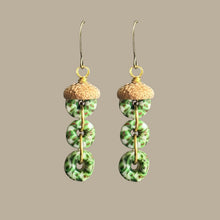 Load image into Gallery viewer, A pair of dangling earrings with gold-tone fishhook wires, small cork-look top pieces, and three stacked green marbled circular beads on each earring, shown against a plain warm gray background.