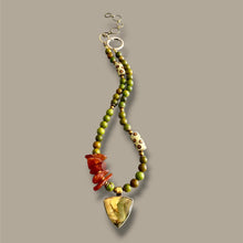 Load image into Gallery viewer, A beaded necklace with green and brown beads, a chunky amber-orange bead cluster, white beads with brown spots, and a large shield-shaped yellow stone pendant, shown against a solid taupe background.