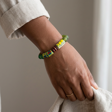 Load image into Gallery viewer, A close-up of a person's hand and wrist wearing a colorful beaded bracelet. The bracelet features green, yellow, red, and white patterned beads, worn over a light beige sleeve.