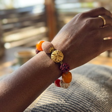 Load image into Gallery viewer, A close-up of a person's wrist wearing a beaded bracelet with orange beads, a pink striped bead, a gold-toned carved bead, and a red knotted accent, with a gold ring on the other hand visible in the background.