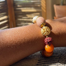 Load image into Gallery viewer, A close-up of a beaded bracelet worn on a person's wrist. The bracelet features a light pink wrapped bead, a round gold-toned decorative charm, a red textured bead, and two smooth orange beads.