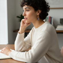 Load image into Gallery viewer, Woman sitting at a desk with a notebook, wearing a white sweater and blue earrings.