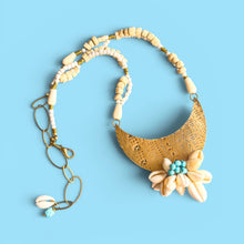 Load image into Gallery viewer, A bohemian-style necklace featuring a large, textured gold crescent moon pendant adorned with a cream flower and turquoise bead center, connected to a beaded chain of cream and white beads with gold spacer beads and a shell charm, set against a solid light blue background.