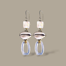 Load image into Gallery viewer, A pair of handmade dangle earrings featuring iridescent square beads, cowrie shells, and clear textured oval drops on silver hooks, set against a solid taupe background.