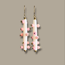 Load image into Gallery viewer, A pair of dangling earrings with vertical white cylindrical beads, small pink beads, and gold-tone hooks, shown against a solid taupe background.