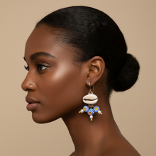 Load image into Gallery viewer, A woman in profile wearing a dangling earring with a white cowrie shell and blue floral charms, set against a solid beige background.