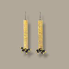 Load image into Gallery viewer, A pair of long rectangular brass earrings with textured patterns and small black  bead clusters at the bottom, shown against a solid taupe background.