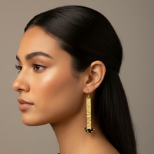 Load image into Gallery viewer, A woman in profile wearing a long, gold-toned drop earring with a vertical bar design and small dark bead accents, against a plain taupe background.
