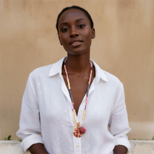 Load image into Gallery viewer, A woman wearing a white button-up shirt and a long beaded necklace with a colorful pendant, standing against a warm beige wall.