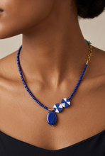 Load image into Gallery viewer, A close-up of a model wearing a beaded necklace with a blue stone pendant and two white-and-blue geometric beads, accented by gold-tone hardware.