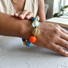 Load image into Gallery viewer, A close-up of a person's wrist and hand wearing a colorful beaded bracelet with turquoise, light green, orange, and gold-toned beads, set against a soft-focus indoor background.